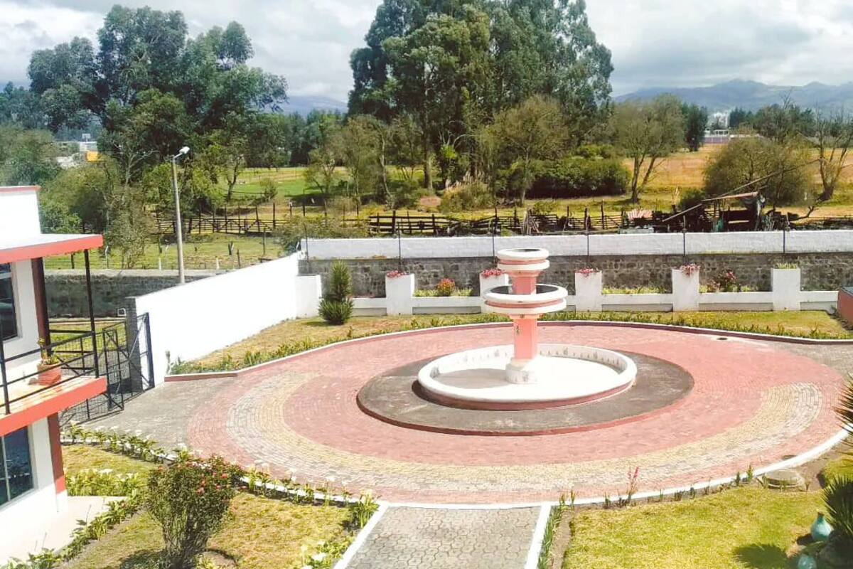 Um caminho de tijolos em formato circular com uma fonte central, cercado por um muro baixo e vegetação.