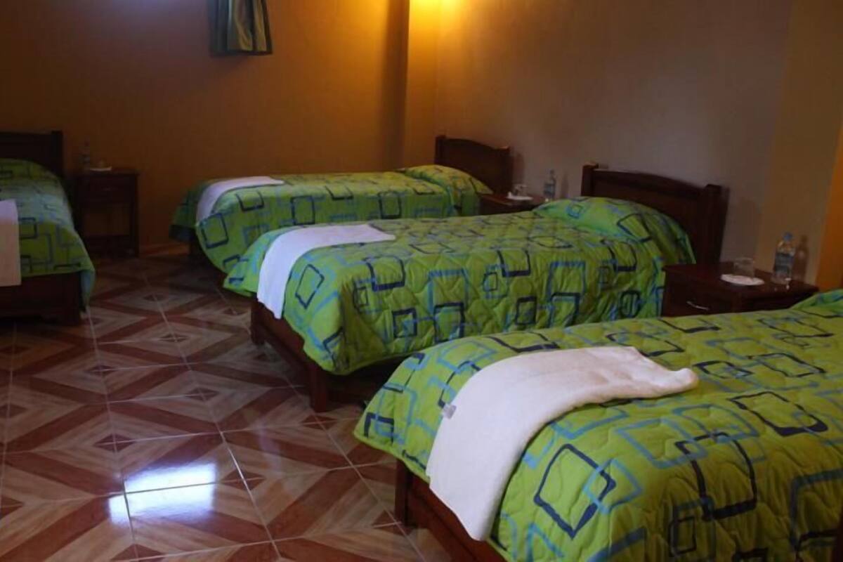 Quarto de hotel com três camas, cada uma com roupa de cama verde e um travesseiro branco. O quarto tem soalho em mosaico e uma pequena mesa com uma garrafa e copos.