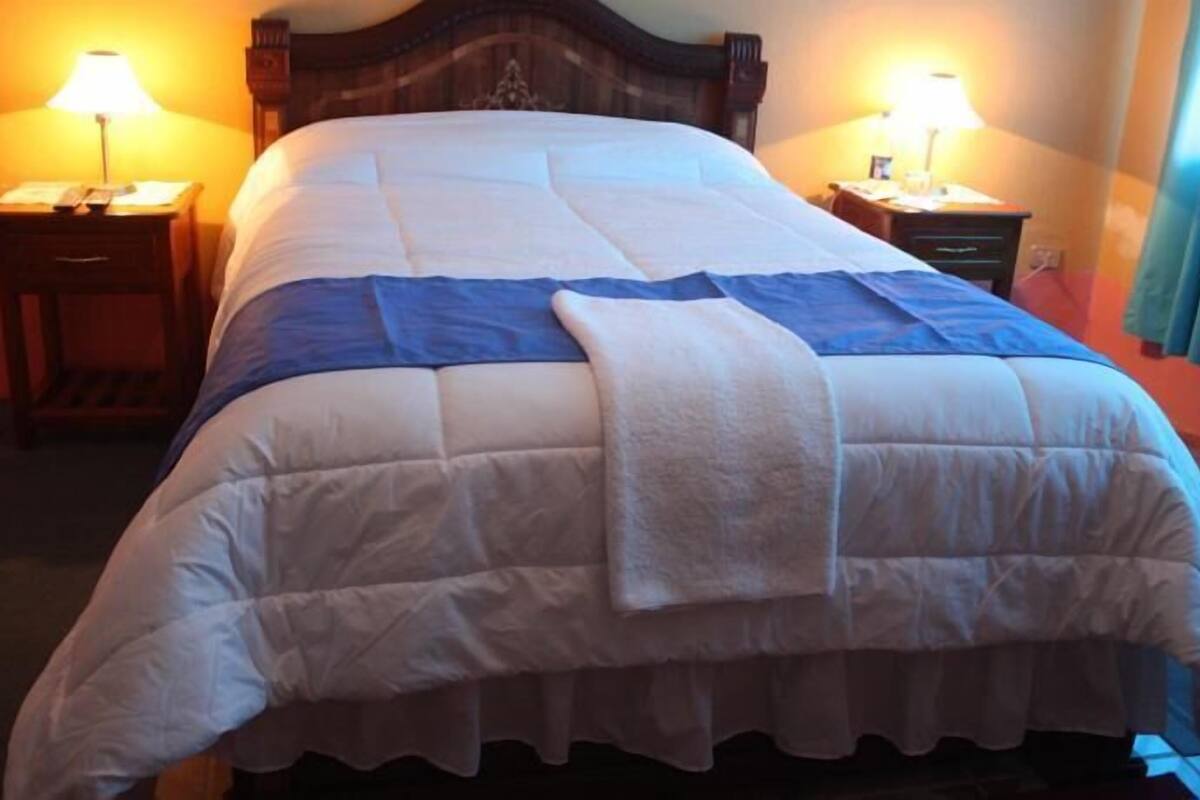 Cama bem arrumada com edredom azul e branco, duas luminárias de mesa e cabeceira de madeira.