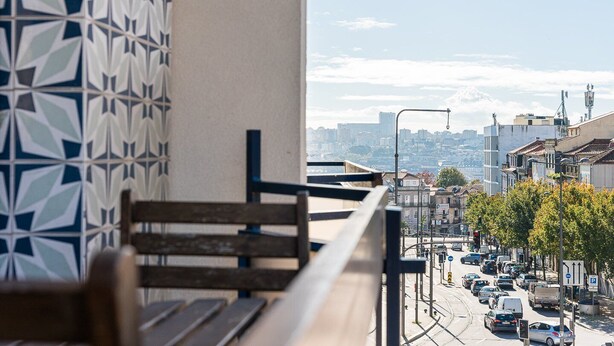A balcony with a patterned wall, a wooden bench, and a view of a busy street with buildings and vehicles. - GuestReady - Cozy Bright apartment with a Balcony (Porto)