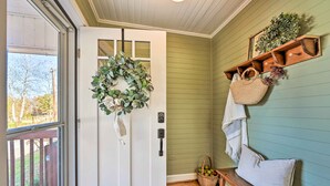 Una puerta con un adorno de laurel, un banco con cojines, una cesta y un estante de madera con una planta y un bolso tejido.