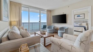 A spacious living room with a large sofa, two armchairs, a coffee table, and a flat-screen TV. A balcony with a view of the sea is visible through the glass doors.