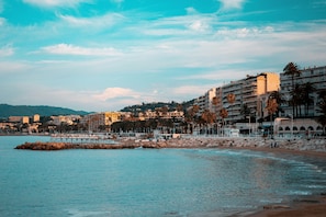 A coastal cityscape with a rocky breakwater, beachfront buildings, and palm trees. - Piscine Rooftop | Chambre +Terrasse Privée (Le Cannet)