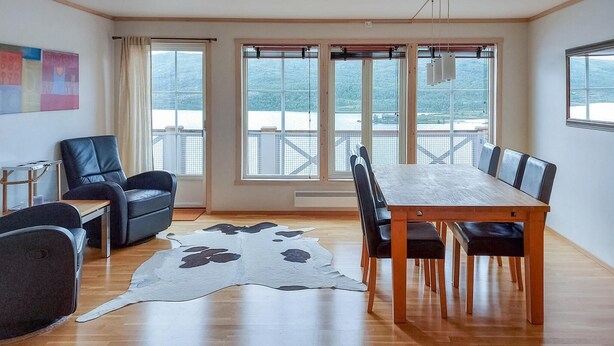A spacious room with a wooden dining table, black leather chairs, a cowhide rug, and large windows offering a view of a lake and mountains.