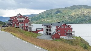 A row of red buildings with balconies, a road in the foreground, and a lake in the background.
