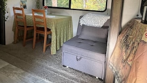 A compact RV interior with a dining area, a bed, and a window with a view of trees. - Quiet, majestic views of the Shire in a cozy glamping set up!  (Brush Prairie)