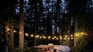 A camper parked on a gravel path surrounded by tall trees, with string lights illuminating the area. - Quiet, majestic views of the Shire in a cozy glamping set up!  (Brush Prairie)