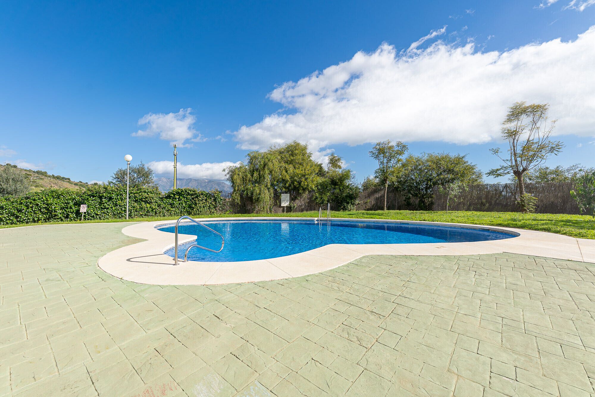 Piscina com escada de metal, rodeada por um espaço pavimentado e vegetação.