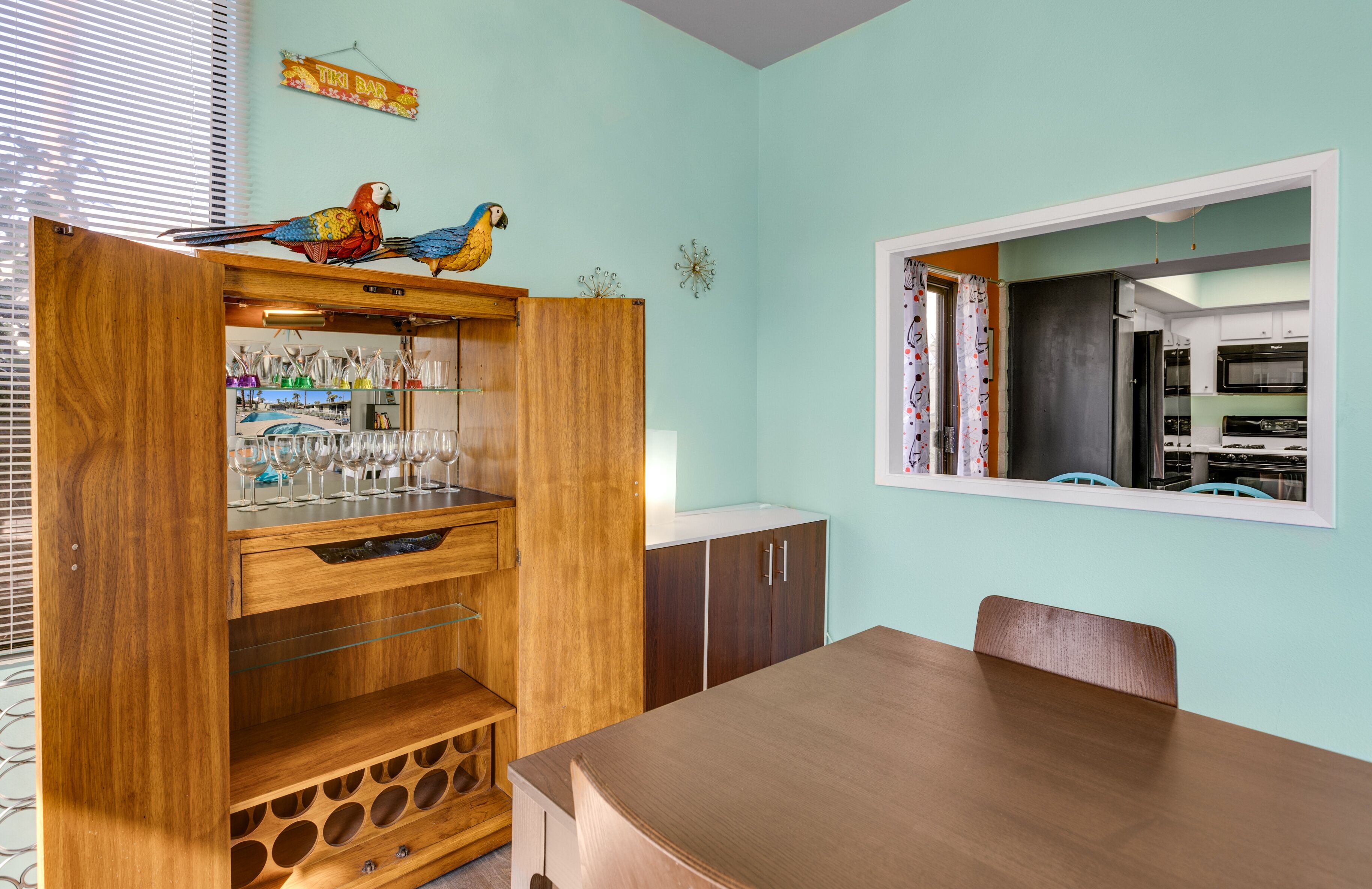 Un coin repas avec une table et des chaises en bois, un buffet en bois contenant des verres à vin et un miroir mural qui reflète l’espace cuisine.