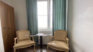 Une pièce avec deux fauteuils beiges, une petite table, une fenêtre avec des rideaux et un radiateur.