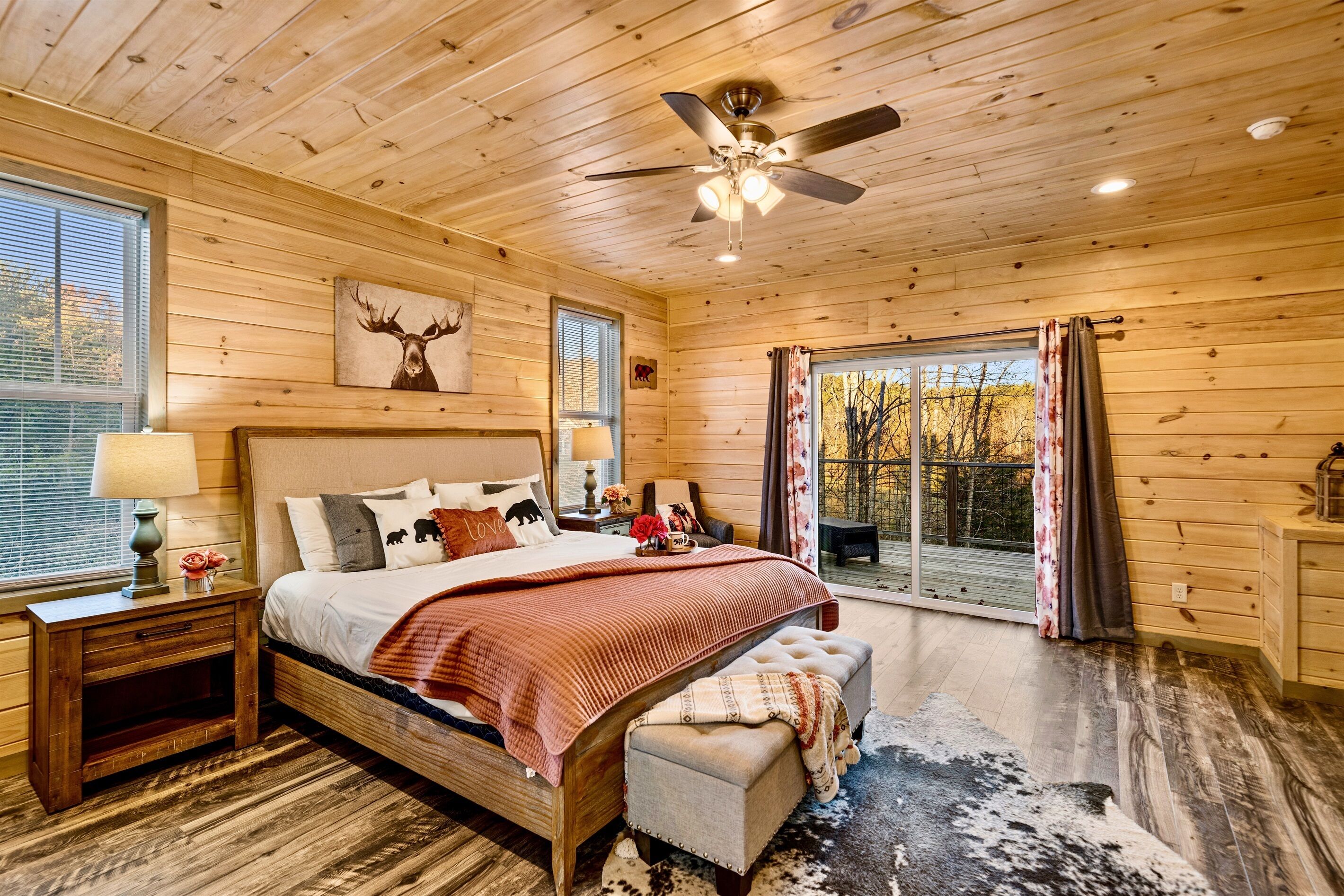 A wooden cabin bedroom with a large bed, a ceiling fan, and a view of the outdoors.