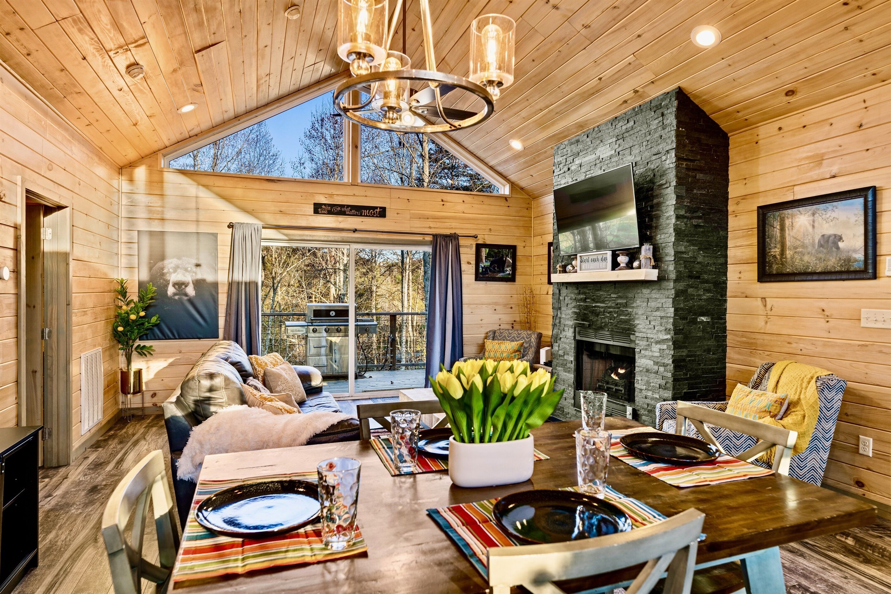 Een gezellig interieur van een blokhut met een eethoek, houten wanden, een open haard en een groot raam met uitzicht op een bos.