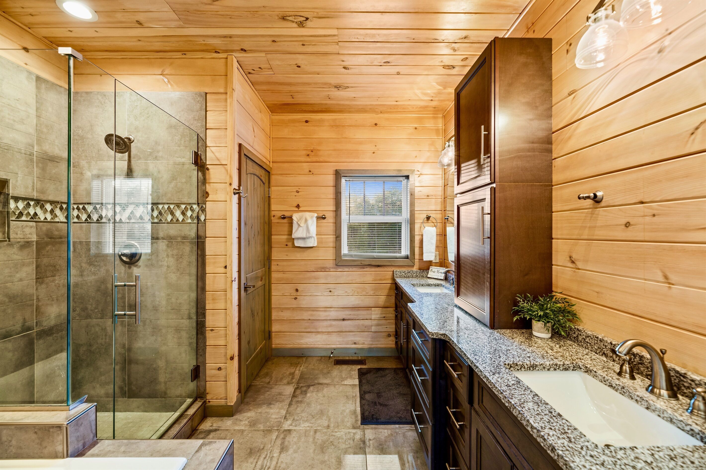 Baño de cabaña de madera con ducha de vidrio, lavamanos y ventana.