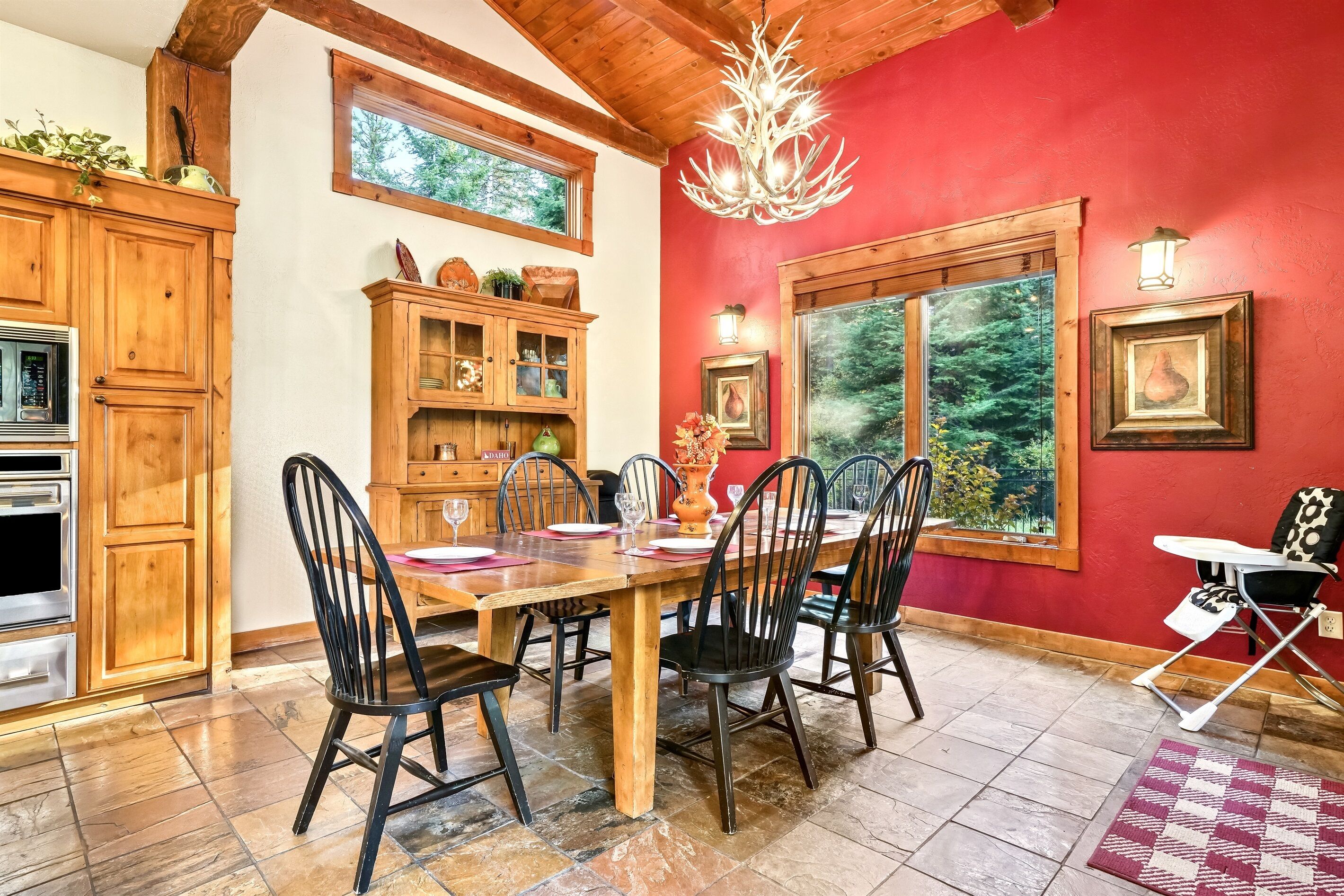 Un coin repas avec une table en bois, des chaises noires, un lustre et un mur rouge.