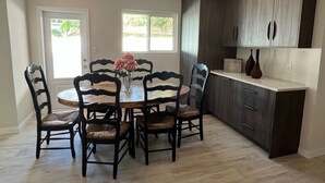 Un coin repas avec une table ronde, six chaises et un vase de fleurs. Il y a deux fenêtres avec des rideaux, un coin cuisine avec des meubles sombres et un comptoir sur lequel sont posés deux vases.
