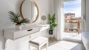 Ein modernes Schlafzimmer mit einem weißen Schminktisch, einem runden Spiegel, zwei Blumentöpfen, einem kleinen Hocker und Blick auf Gebäude durch ein großes Fenster.