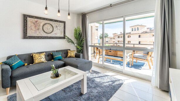 Ein modernes Wohnzimmer mit grauem Sofa, einem Glastisch und einem großen Fenster mit Blick auf Gebäude und einen Pool.