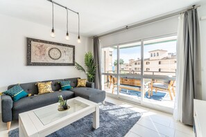 Ein modernes Wohnzimmer mit grauem Sofa, einem Glastisch und einem großen Fenster mit Blick auf Gebäude und einen Pool.