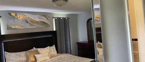 A bedroom with a large mirror, a bed with multiple pillows, a dresser, and a painting on the wall.