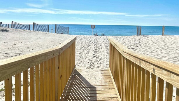 Une promenade en bois mène à une plage de sable fin, sous un ciel bleu clair et une mer calme.