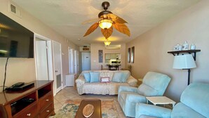 Un salon avec un ventilateur de plafond, un téléviseur, un canapé, une table basse et une petite table.