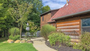 Une cabane en rondins avec un chemin de gravier menant à l’entrée, entourée d’un jardin bien entretenu où poussent différentes espèces de plantes et où se trouve un banc.