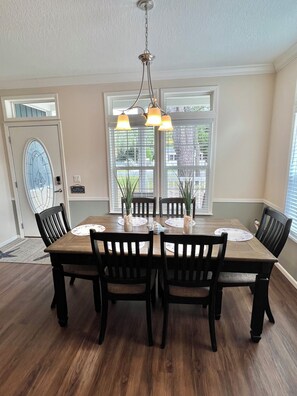 A dining room with a wooden table, four chairs, a chandelier, and large windows.