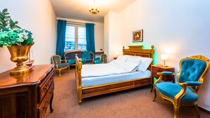 Une chambre d’hôtel avec un lit en bois, un fauteuil bleu, un bureau avec une lampe et une vue sur la ville par la fenêtre.