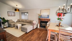 Ein Wohnzimmer mit Ecksofa, Kamin, Esstisch mit Stühlen und einer Pendelleuchte.