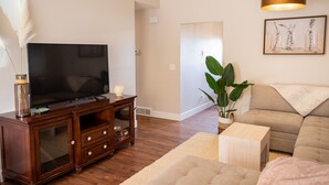 Ein Wohnzimmer mit Flachbildfernseher auf einer Holz-Unterhaltungseinheit, einem beigefarbenen Sofa, einem Couchtisch und einer großen Topfpflanze.