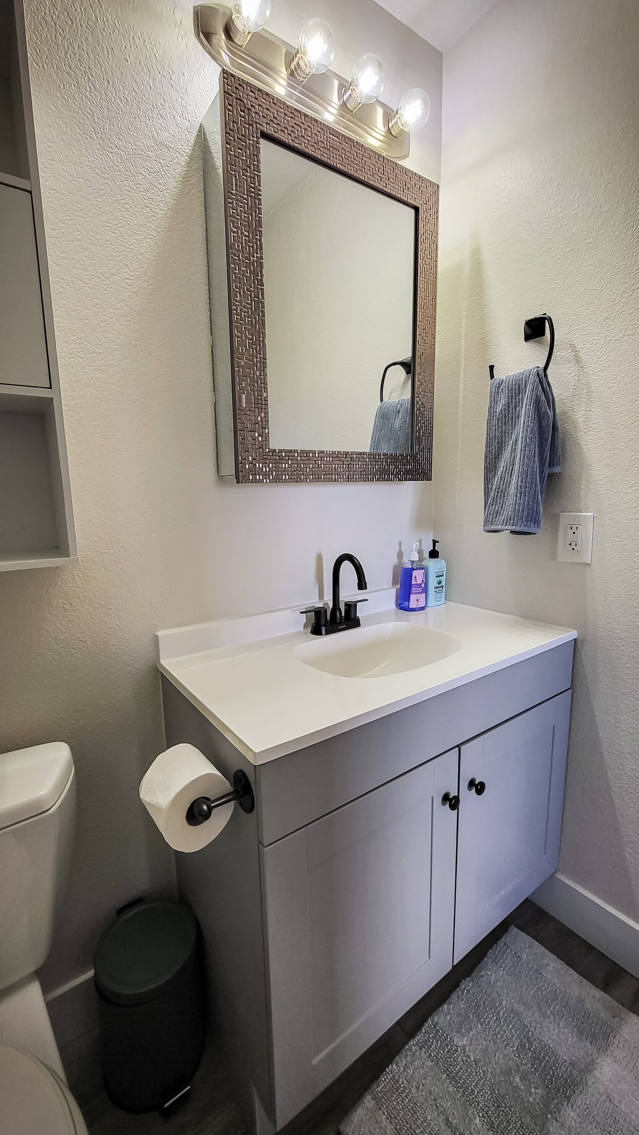 A bathroom with a white sink, a mirror with lights, a towel rack, and toiletries.