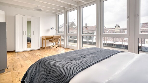 A bedroom with a large bed, a desk, and a wardrobe. There are large windows offering a view of buildings outside.