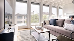 A modern living room with a sofa, coffee table, and TV. Large windows offer a view of the city.