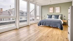 A spacious bedroom with large windows, a wooden floor, a bed with a blue and white checkered bedspread, and two framed pictures on the wall.