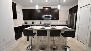 A modern kitchen with dark cabinets, a central island with bar stools, stainless steel appliances, and pendant lighting. - 5BR/5BA Game/Pool Villa 9 Miles to Disney (Kissimmee)