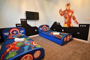 A room with two beds, each with superhero-themed bedding. There is a flat-screen TV mounted on the wall.