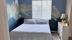 Quarto com cama, janela com persianas, mesa de cabeceira com abajur e cômoda.