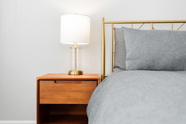 A neatly made bed with a grey blanket and pillows, a wooden nightstand with a lamp, and a gold-framed headboard.