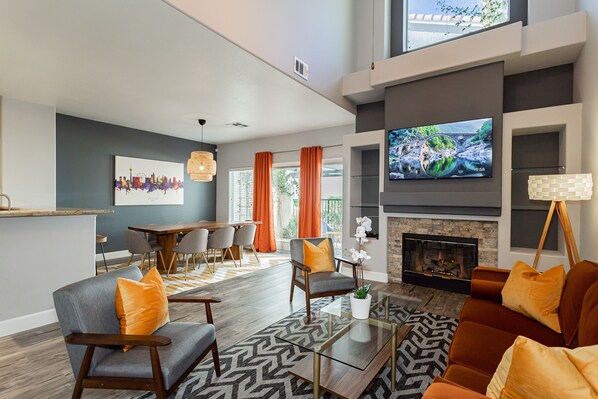 A modern living room with a fireplace, a television, and a dining area with orange curtains.