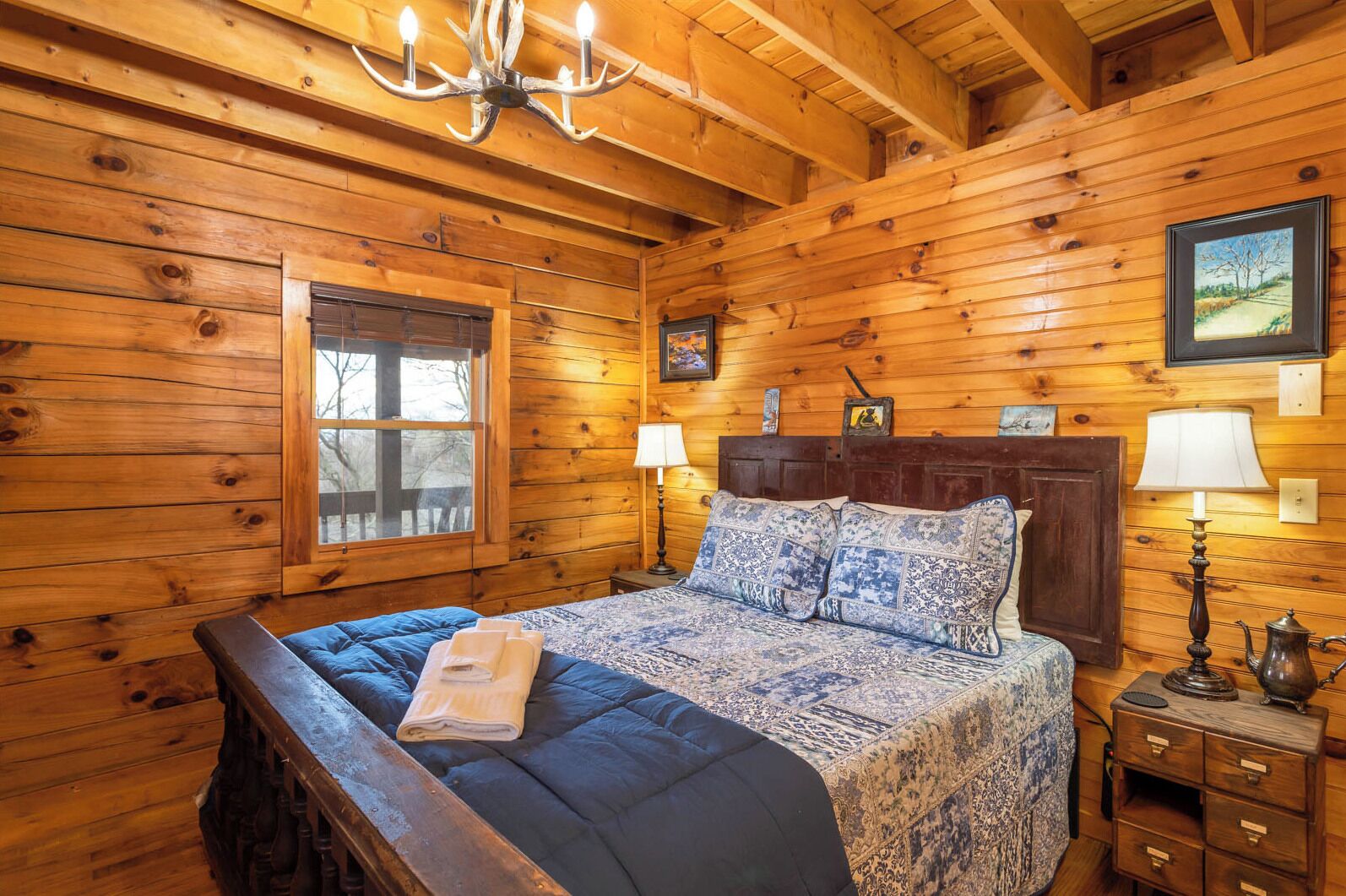 A wooden cabin bedroom with a bed, two bedside lamps, a chandelier, a window, and a picture on the wall.