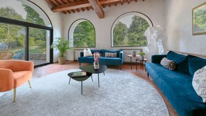 Ein modernes Wohnzimmer mit einem blauen Sofa, einem orangefarbenen Sessel und einem Couchtisch. Es gibt große Fenster mit Rundbögen, eine Holzbalkendecke und eine Statue auf einem kleinen Tisch.