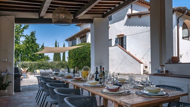 Ein Essbereich im Freien mit einem langen Holztisch, gedeckt für eine Mahlzeit, umgeben von Stühlen, unter einer Pergola mit einer hängenden Leuchte.