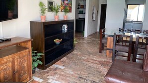 Ein Wohnzimmer mit Fernseher, Holzschrank und Topfpflanzen. Es gibt eine Essecke mit Tisch und Stühlen.