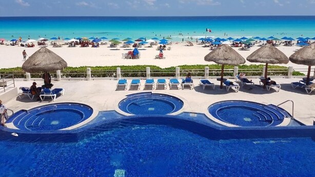 Ein Poolbereich mit mehreren Schwimmbecken und Liegestühlen, ein Strand mit Sonnenschirmen und Menschen sowie ein klarer, blauer Himmel.