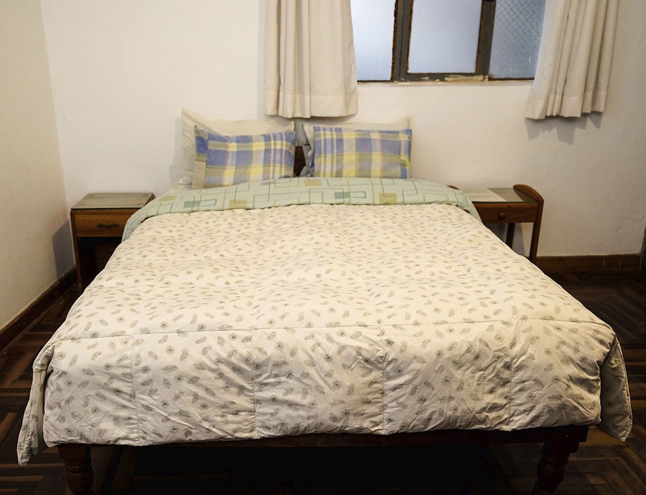 Quarto com cama, duas mesas de cabeceira, janela com cortinas e piso de madeira.