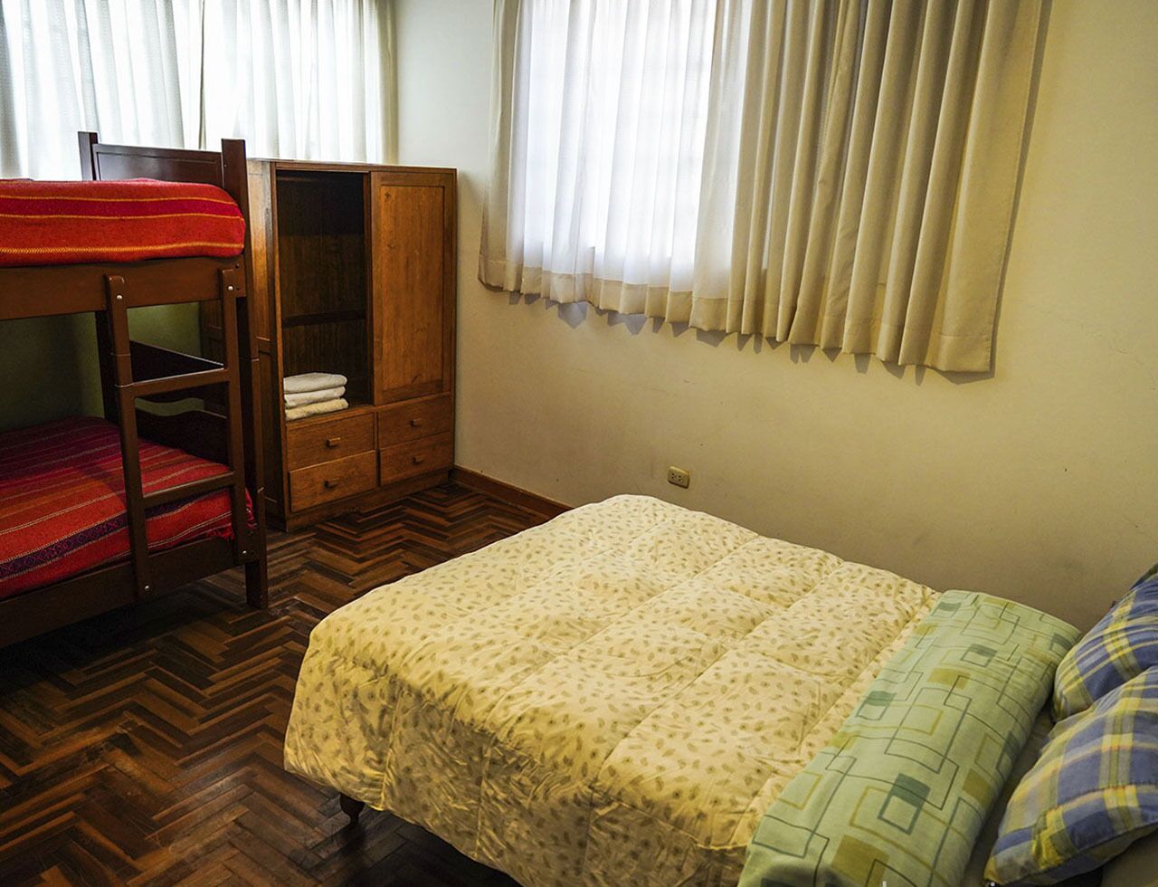 A bedroom with a bunk bed, a single bed, a wooden wardrobe, and a window with curtains.