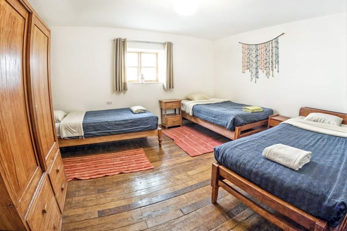 A dormitory room with three wooden beds, a wooden wardrobe, a window with curtains, and a wall hanging.