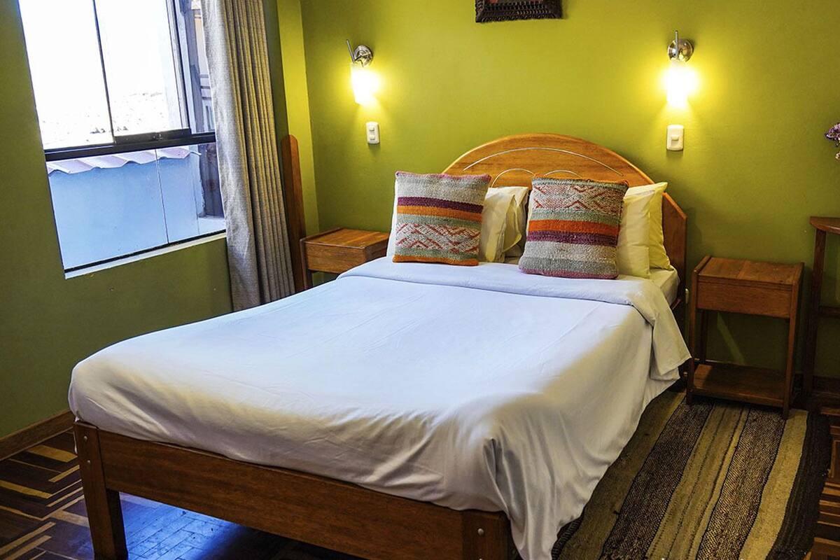 A neatly made bed in a room with green walls, two bedside tables, and a window with curtains.