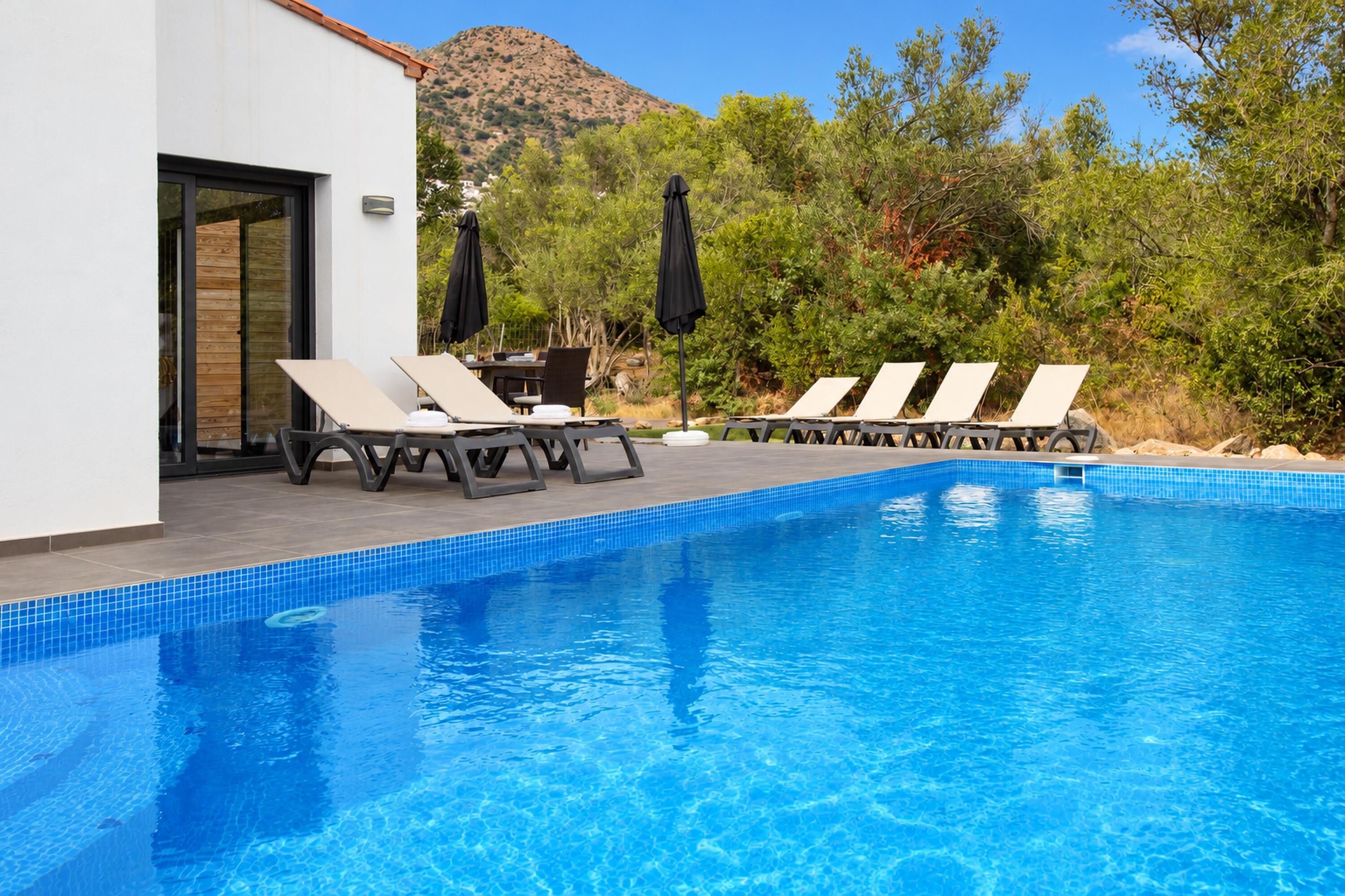 A swimming pool with lounge chairs and umbrellas, surrounded by greenery and a mountain in the background.