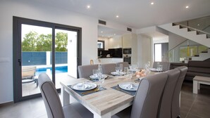 A modern dining area with a wooden table set for six, surrounded by grey upholstered chairs. A glass door leads to an outdoor seating area with a pool. - Villa for rent in Mas Busca, Roses, Costa Brava: Spacious villa with private pool, ideal for families, 3 km from Roses beach. (Roses)